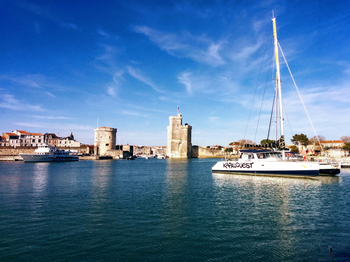 Château médiéval, voilier, port animé, ciel bleu, paysage côtier.