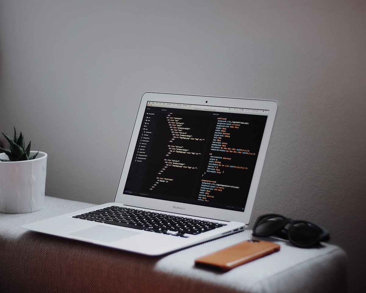 Ordinateur portable ouvert avec code de programmation à l'écran, sur un bureau avec une plante et des lunettes.