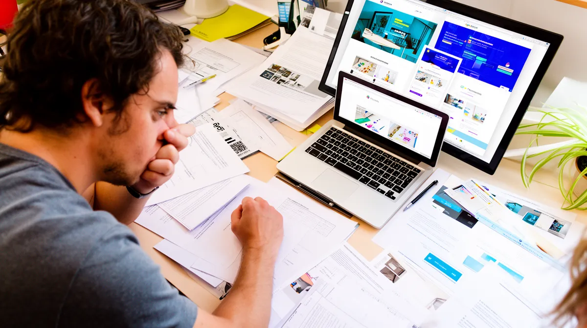 Homme concentré sur son travail, entouré d'ordinateurs et de documents.